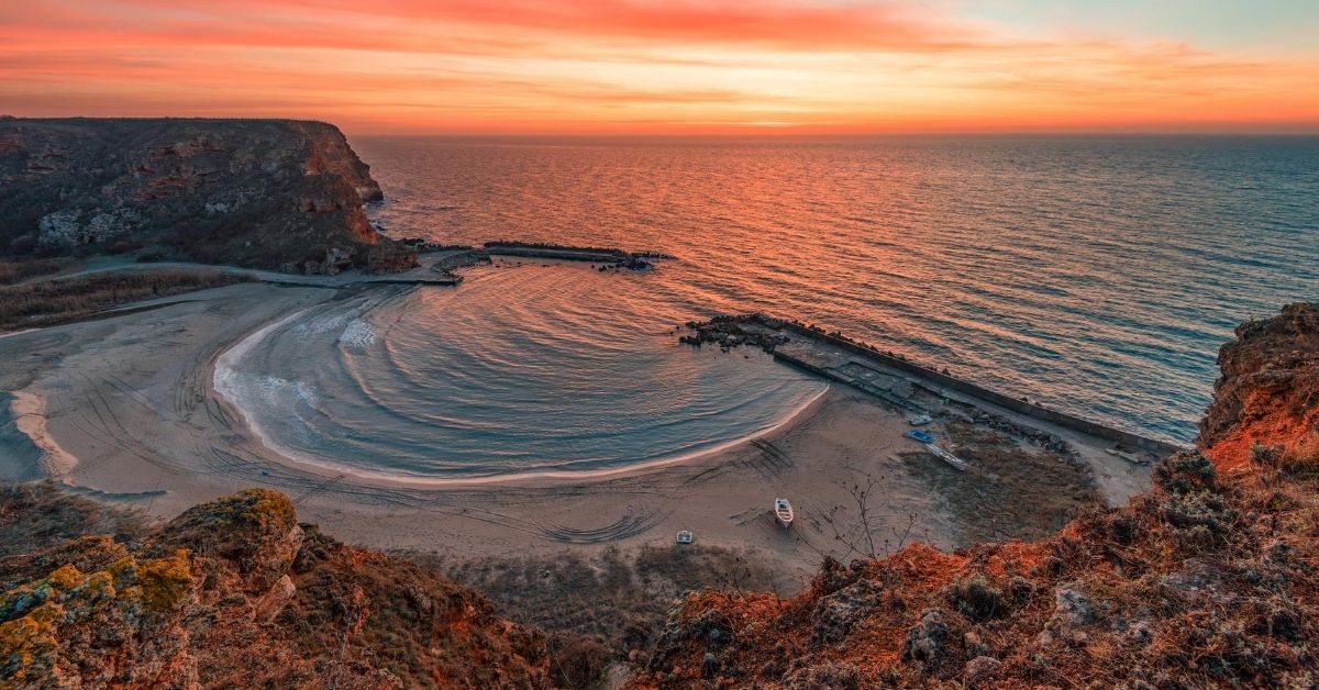 beautiful round beach with boats on sand at Bolata Bay surrounded by cliffs with orange skies at dusk