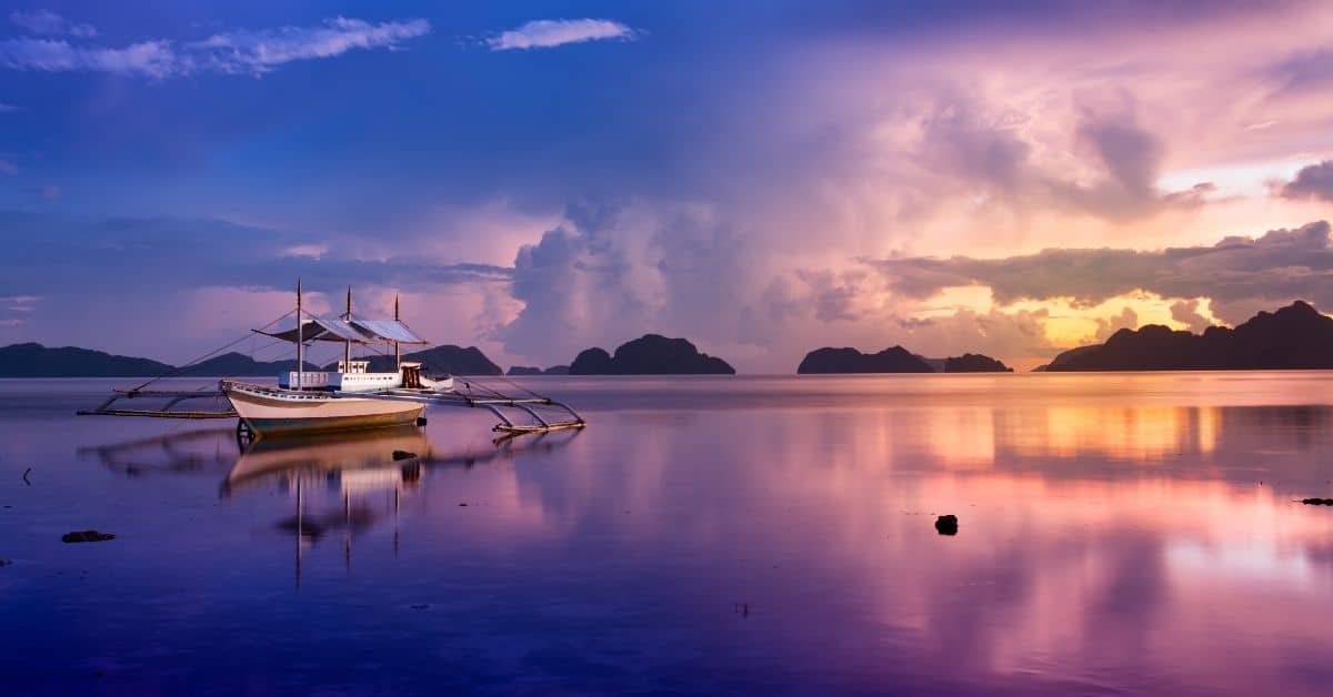 beautiful purple and blue sunset reflected in water with boat and surrounding mountains in Palawan, Philippines