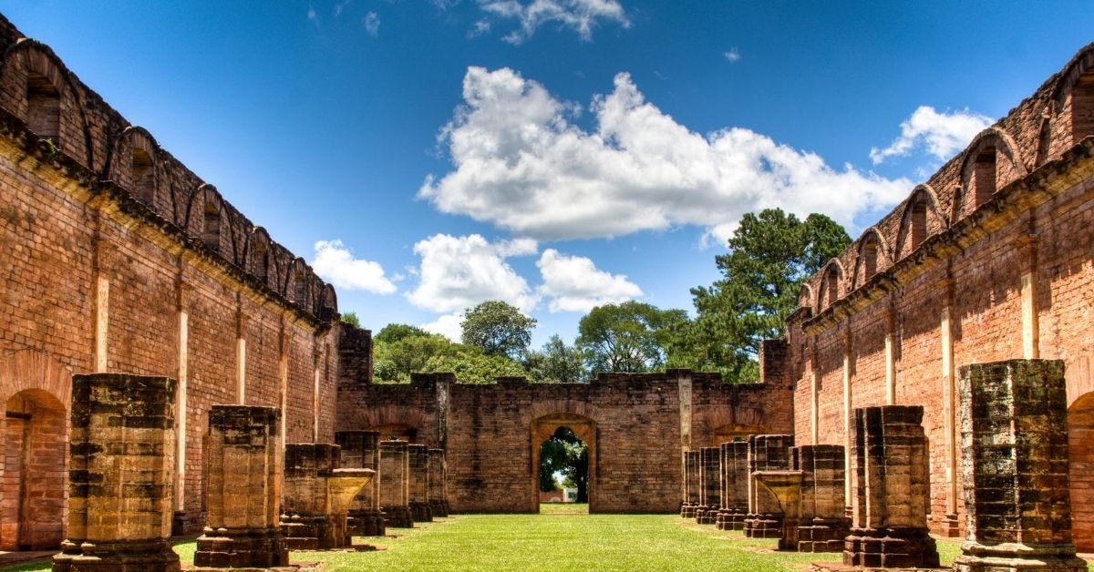 red brick ruins with columns and tall walls with trees and cloudy blue skies