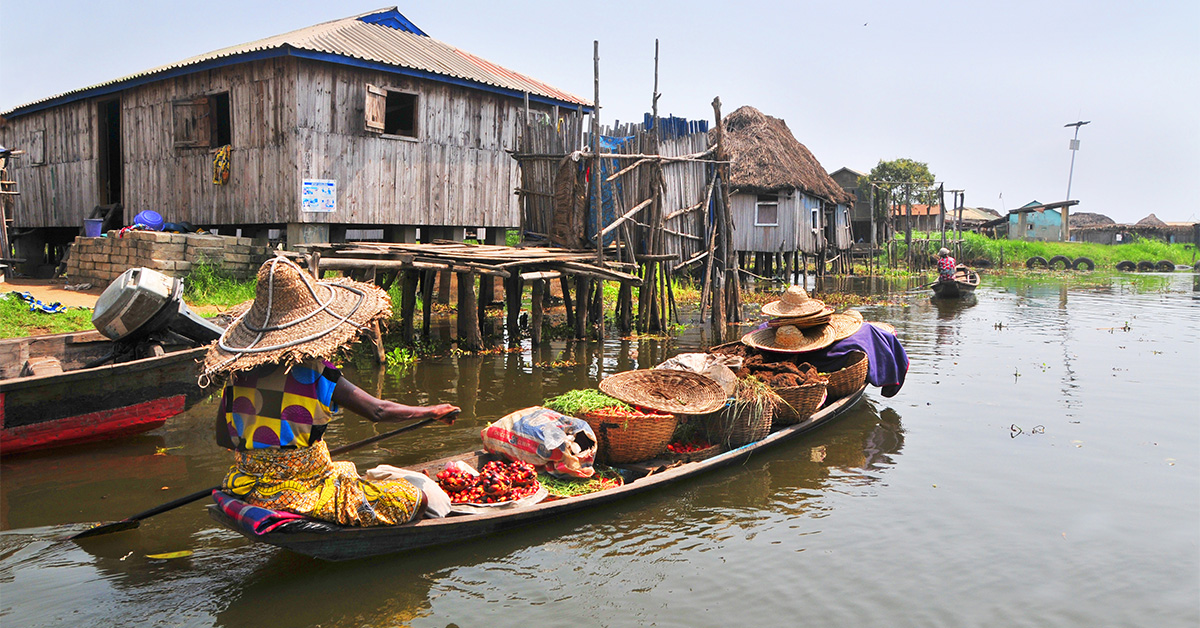 In Benin, a woman in small wooden canoe with colorful food rows on lake with stilt houses in Ganvie