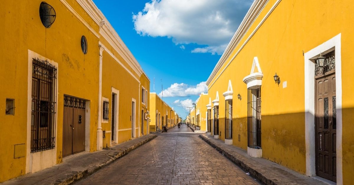 colonial facades of houses along each side of a brick street, all painted yellow with blue skies and white puffy clouds