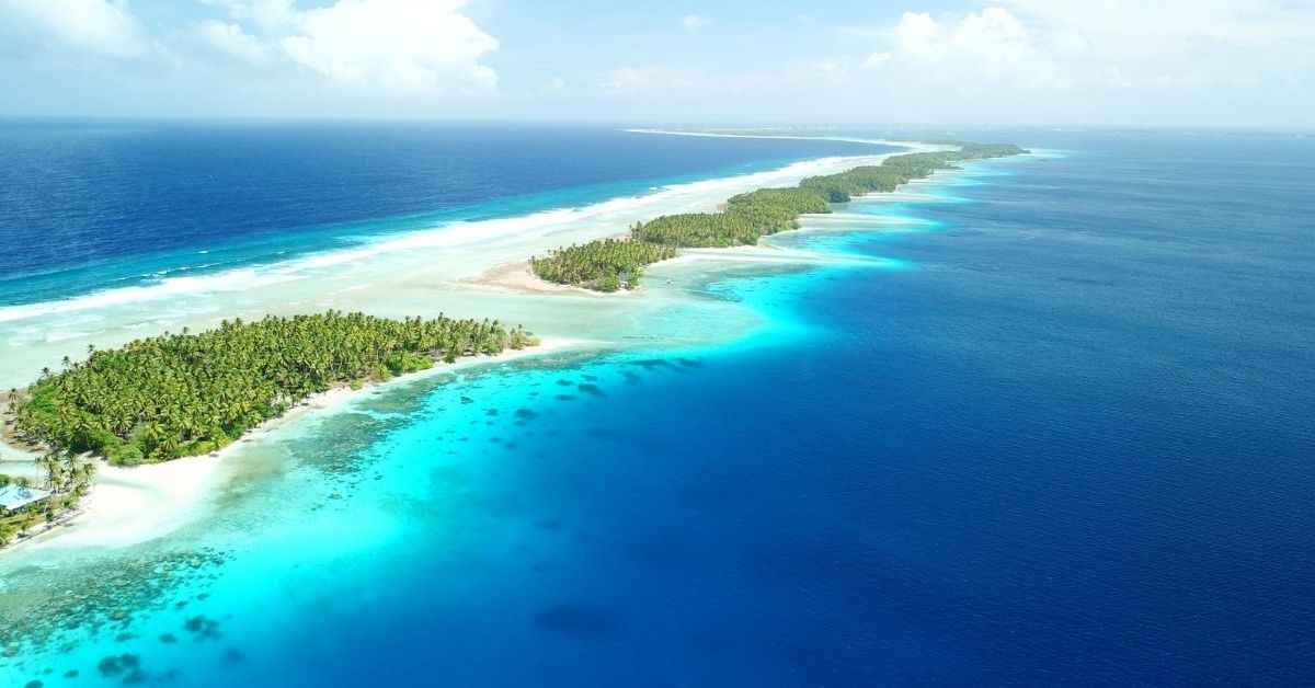 aerial view a chain of palm tree covered atolls with white sand beaches surrounded by the Pacific in shades of blue
