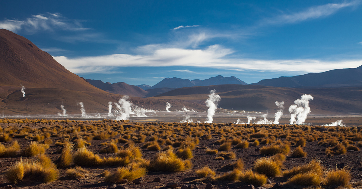 brown toned landscape of white steam rising from the ground or geothermal activity in the Atacama with hills and mountains with blue sky and white clouds