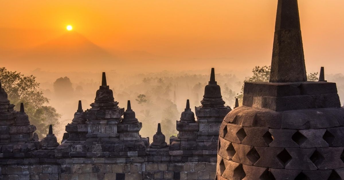 stupas atop the Borobudur temple with the sunrise directly over volcano in the distance overlooking the valley with palm trees in the morning mist