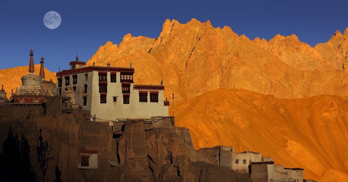 The moon rises against a blue sky with the Lamayuru Monastery perched atop a mountain—facing larger mountains in the distance with the glow of the sun turning them the color of rust.