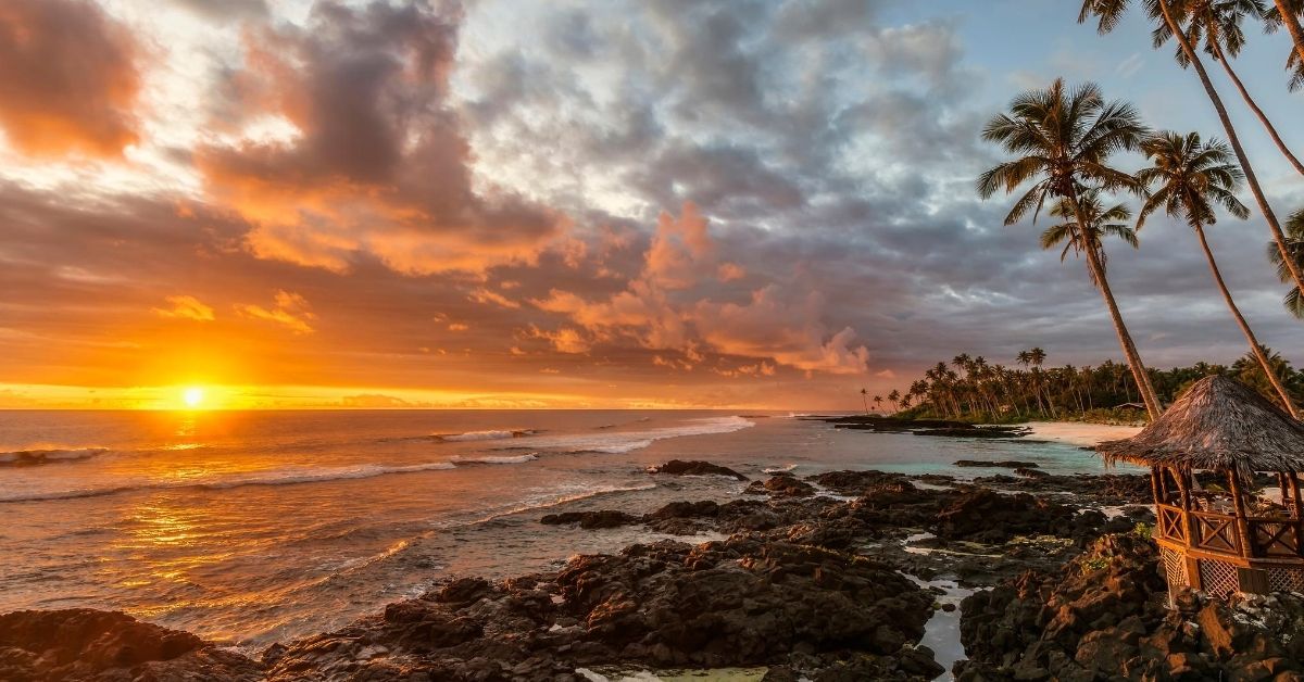 gazebo on coast of samoa with stretch of beach lined with palm trees and a dramatic orange glowing sunset in the clouds