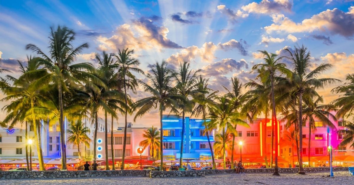 neon lights illuminating buildings with art deco architecture, palm trees and people sitting on stone wall on the beach